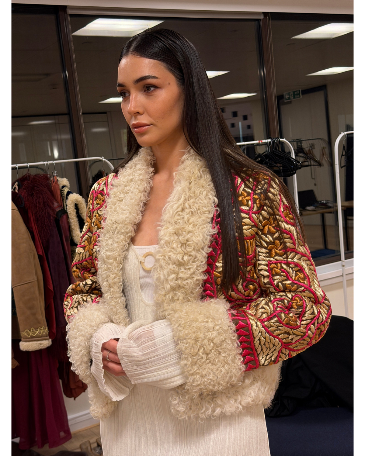 Aql&Nafs handbestickte, kurze afghanische Jacke mit gold-pinkem Muster und cremefarbenem Lammfellbesatz an einer Frau.