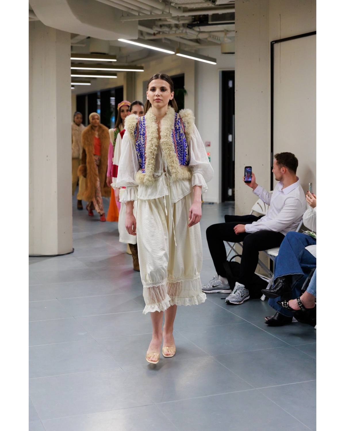 Model auf dem Laufsteg trägt eine Aql&Nafs Nargis Weste mit blauer Stickerei und beigem Lammfellbesatz über weißem Kleid
